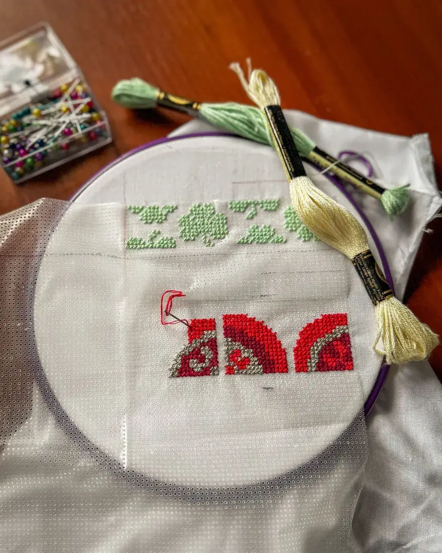 Bastidor con un bordado contado en rojo y verde, junto a madejas de hilo de mouliné y alfileres de colores